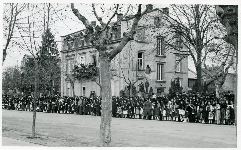 Au bord de l'avenue, la foule est compacte. Les maisons portent encore les stigmates des combats.