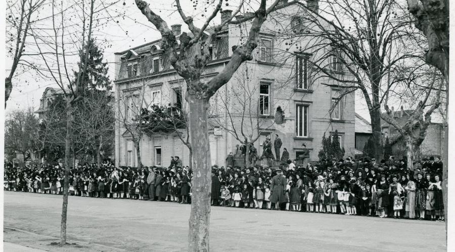 Au bord de l'avenue, la foule est compacte. Les maisons portent encore les stigmates des combats.